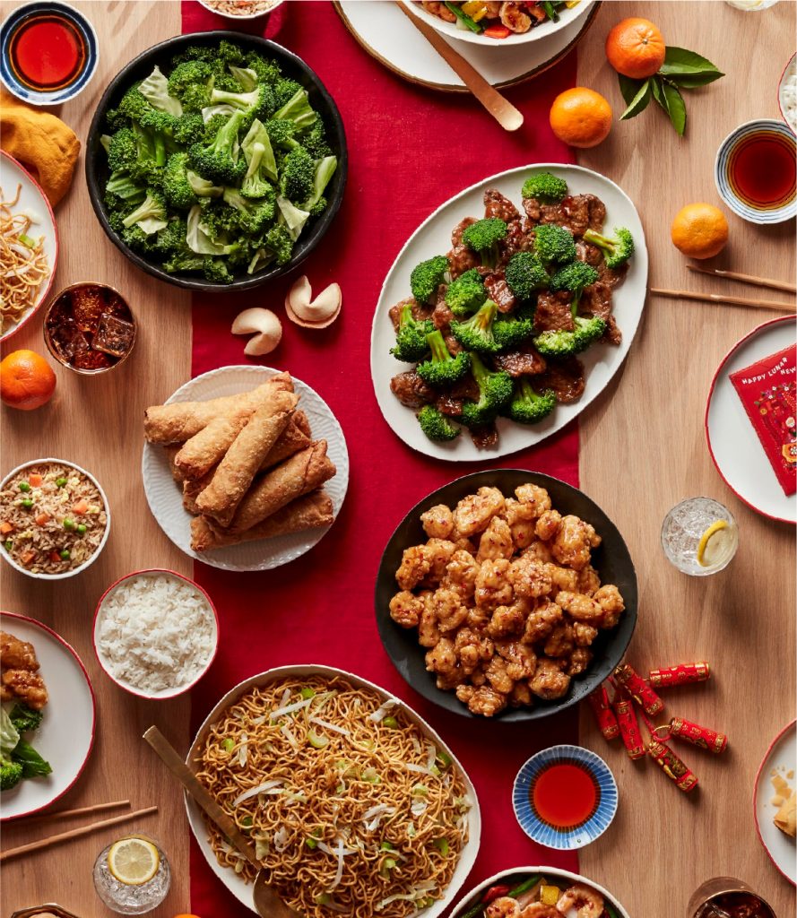Variedad de platos asiáticos incluyendo brócoli salteado, rollitos de primavera, pollo agridulce y fideos, servidos en una mesa festiva decorada con mandarinas y sobres rojos